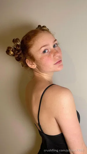 Woman with red hair in braids, wearing black spaghetti strap top, standing indoors against plain