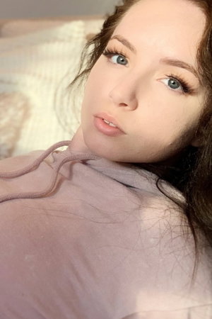 Woman with blue eyes and dark hair wearing a pink hoodie, lying on a bed.