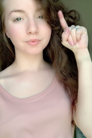 Woman with curly brown hair in pink tank top indoors, making finger gesture.