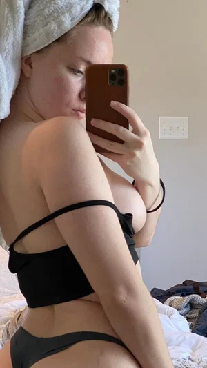 Woman in black lingerie and towel on head, taking mirror selfie in bedroom.