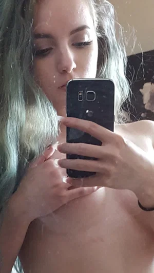 Woman with blue hair taking mirror selfie, bare chest, holding phone.