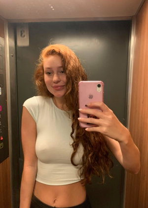 Curly red-haired woman in white crop top taking selfie in elevator.