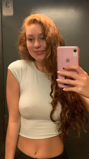 Woman with curly red hair in white crop top and black bottoms, taking mirror selfie indoors.