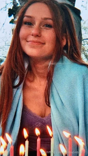 Woman with long brown hair wearing purple top and blue cardigan, holding lit candles outdoors.