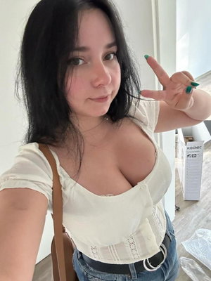 Woman with dark hair in white top and jeans, making peace sign indoors.