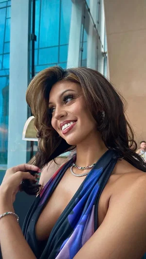 Woman with long brown hair in a colorful halter top, smiling outdoors near a glass building.