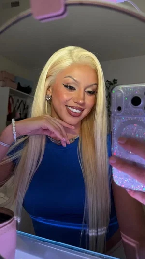 Woman with long blonde hair in blue top taking mirror selfie indoors.