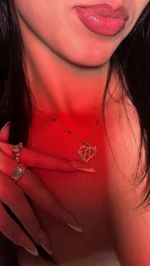 Woman with dark hair and red lips, wearing a heart pendant necklace, hand near chest.