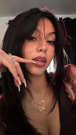 Woman with long dark hair and gold necklace, wearing dark top, posing indoors.