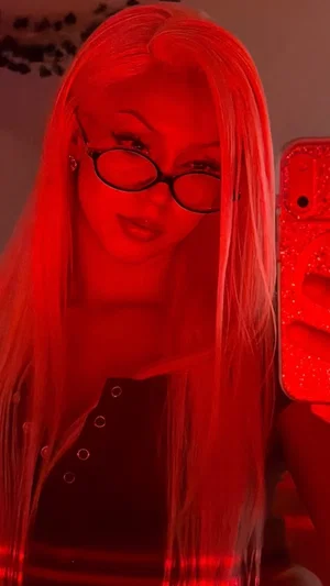 Woman with long blonde hair and glasses takes mirror selfie in red lighting, wearing black top.