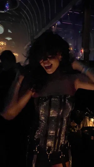 Woman with dark hair in a shiny corset dancing in a dimly lit indoor setting.