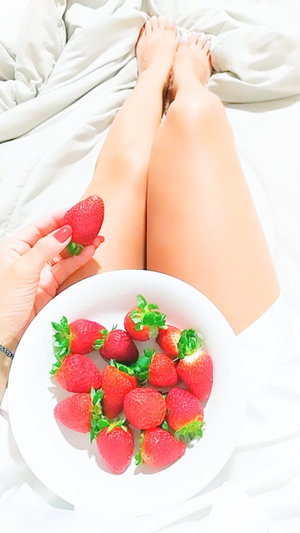 Woman with bare legs holding strawberry, plate of strawberries on bed.