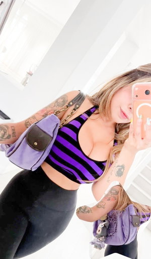 Woman with tattoos wears purple striped crop top and black leggings, holding phone for mirror