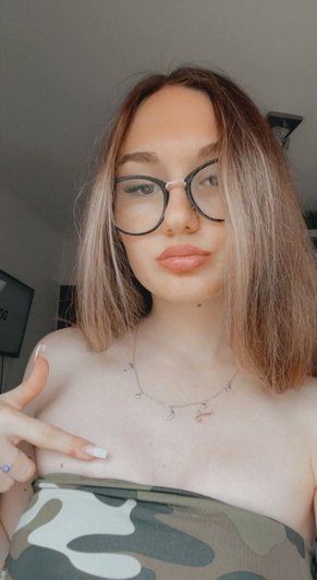 Woman with glasses and shoulder-length hair wearing a camo top, pointing at her chest indoors.