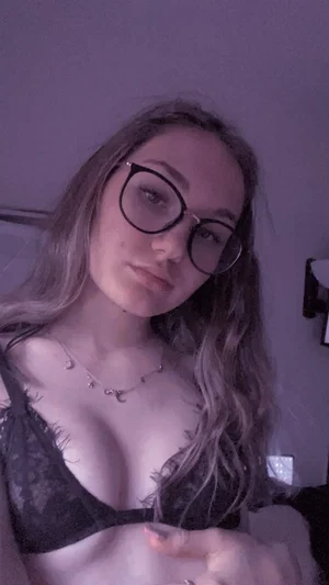 Woman with wavy hair and glasses wearing black lace bra in indoor setting.