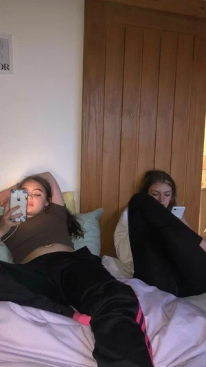Two women lying in bed, one taking a selfie, wearing casual clothes, indoor setting.