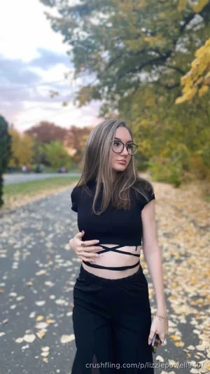 Woman in black crop top and pants standing on leafy path outdoors.