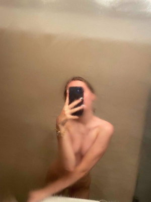 Woman taking mirror selfie in bathroom, topless with arm covering chest.