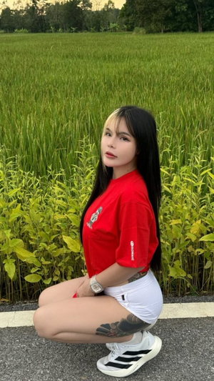 Woman with long black hair in red shirt squatting in green field.