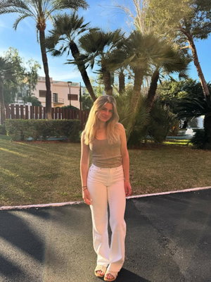 Blonde woman in tank top and white pants standing outdoors near palm trees.