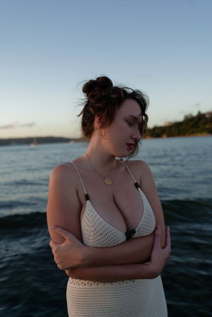 Curvy woman in white crochet dress stands by the water at dusk.