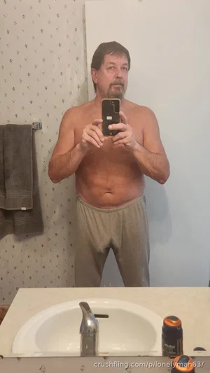 Shirtless man with beard in bathroom mirror selfie, wearing gray sweatpants.