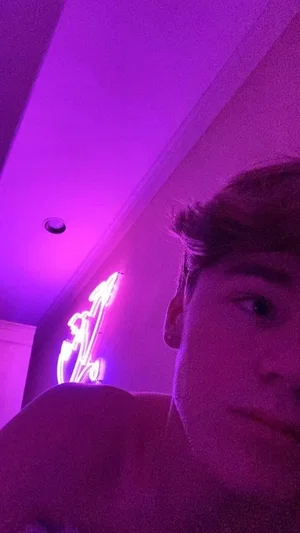 Shirtless man in purple-lit room with neon sign, close-up selfie.