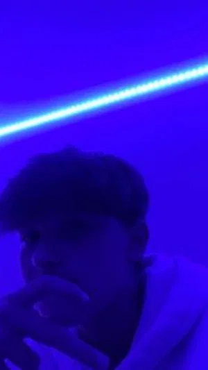 Person in dim blue lighting, wearing light-colored top, hand near face.