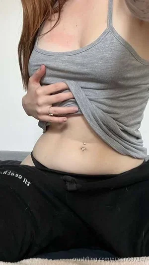Woman in gray tank top and black pants, sitting, pulling top to show midriff.