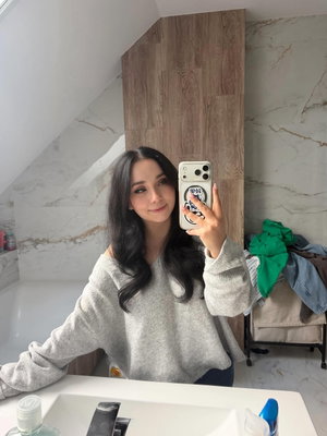 Woman with long dark hair in gray sweater taking mirror selfie in bathroom.