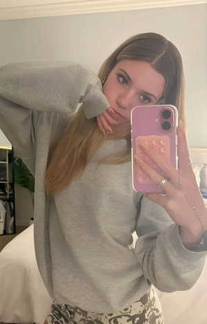 Woman with long blonde hair in grey sweatshirt taking mirror selfie indoors.