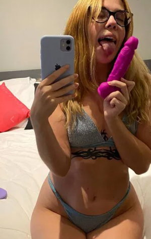 Blonde woman with glasses in bikini taking bedroom selfie.