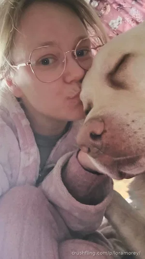 Woman with glasses in pink robe kissing dog indoors.