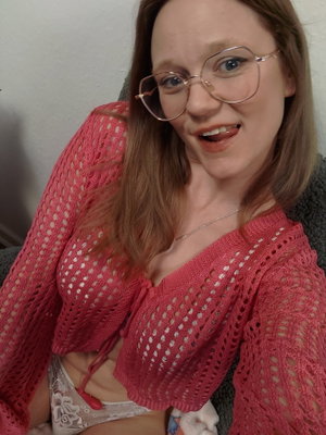 Woman with glasses in pink crochet top and lace underwear, smiling in indoor setting.