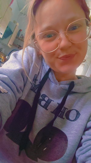 Woman with glasses and blonde hair wearing a gray hoodie, taking a selfie indoors.