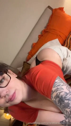 Woman with glasses and arm tattoo in red shirt, lying on bed with orange pillow.