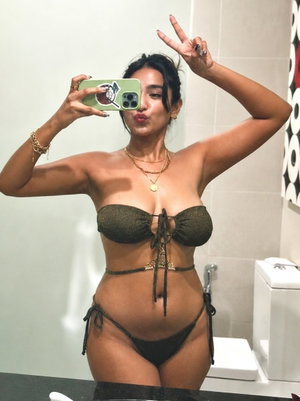 Woman in black bikini taking mirror selfie in bathroom, making peace sign.