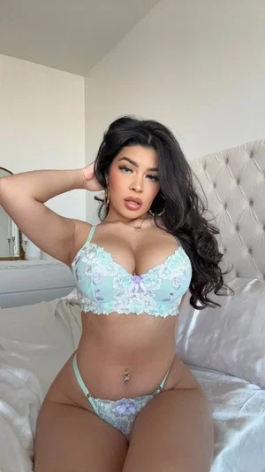 Woman in light blue lace lingerie sitting on bed, dark hair, indoor setting.