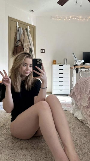 Blonde woman in black bodysuit taking selfie in bedroom.