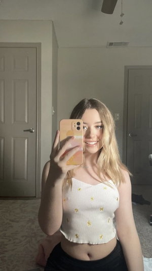 Blonde woman in white crop top taking selfie indoors.