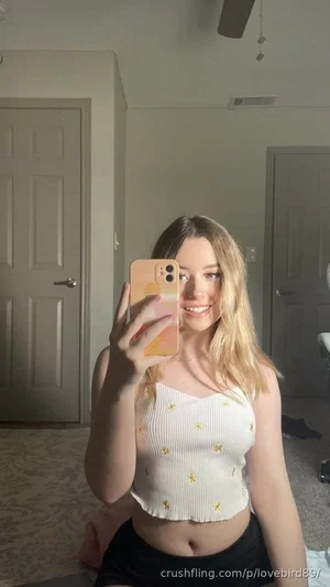 Woman with blonde hair in white crop top and black shorts, sitting indoors taking mirror selfie.