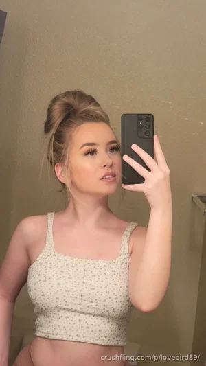 Woman with blonde hair in bun, wearing floral crop top, taking mirror selfie indoors.