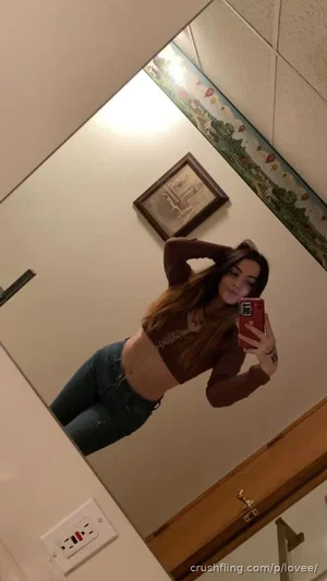 Woman with long dark hair in a brown crop top and jeans takes mirror selfie in a room with framed