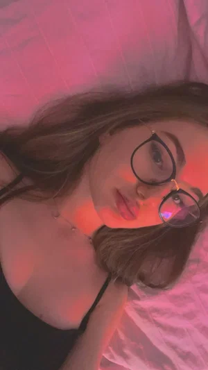 Woman with glasses lying on bed, wearing black top, pink lighting.