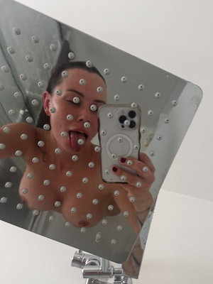 Dark-haired woman taking topless mirror selfie in bathroom.