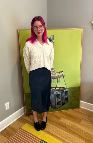 Woman with pink hair in white sweater and black skirt standing indoors near artwork.