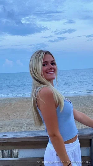 Blonde woman in blue crop top and white pants posing by beach at dusk.