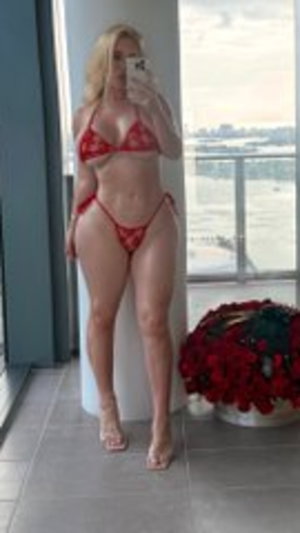 Blonde woman in red lace lingerie posing by window with city view.