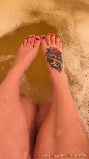 Woman's legs and feet in a bubble bath, one foot with a skull and rose tattoo, red toenails.