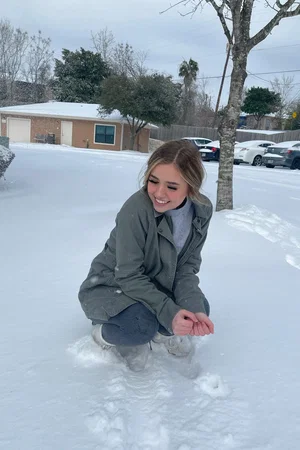 Blonde woman in green jacket and gray pants squats in snow.